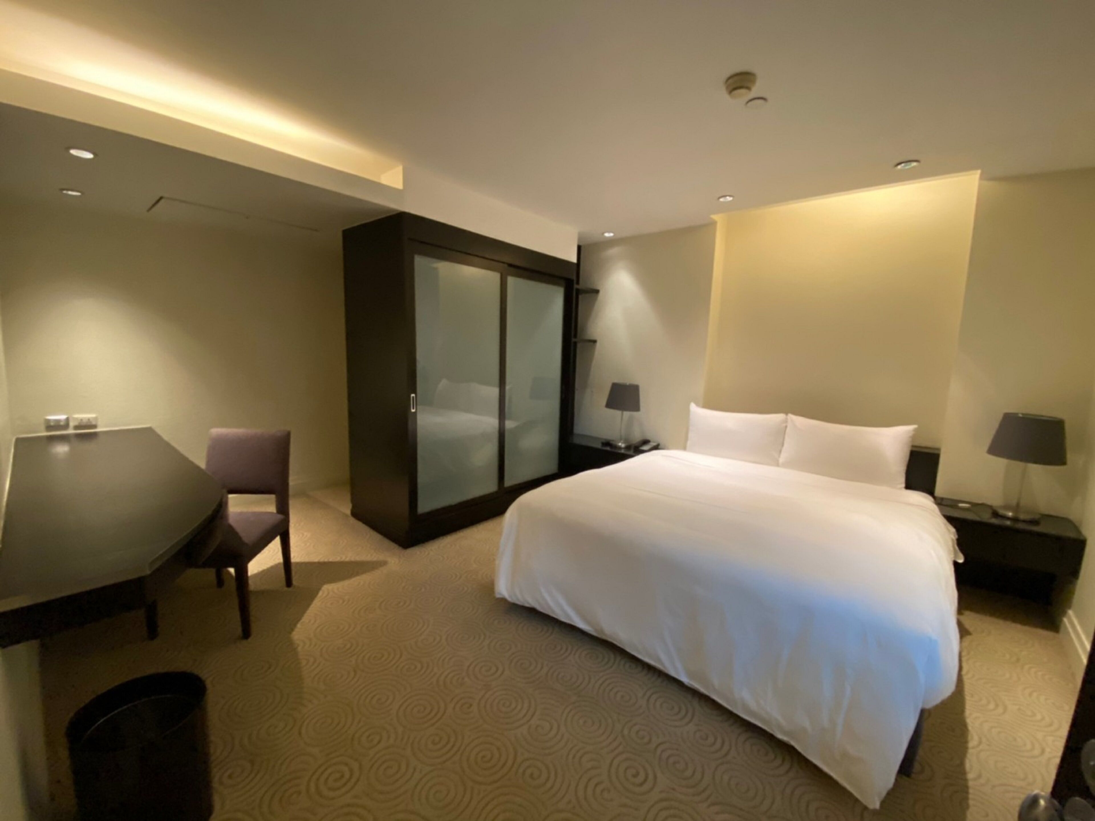 A hotel room with a large bed, a desk, a chair, a wardrobe, and two bedside tables with lamps.