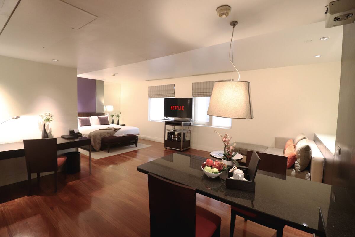 Una habitación de hotel moderna con zona de comedor, cama, televisor y lámpara.
