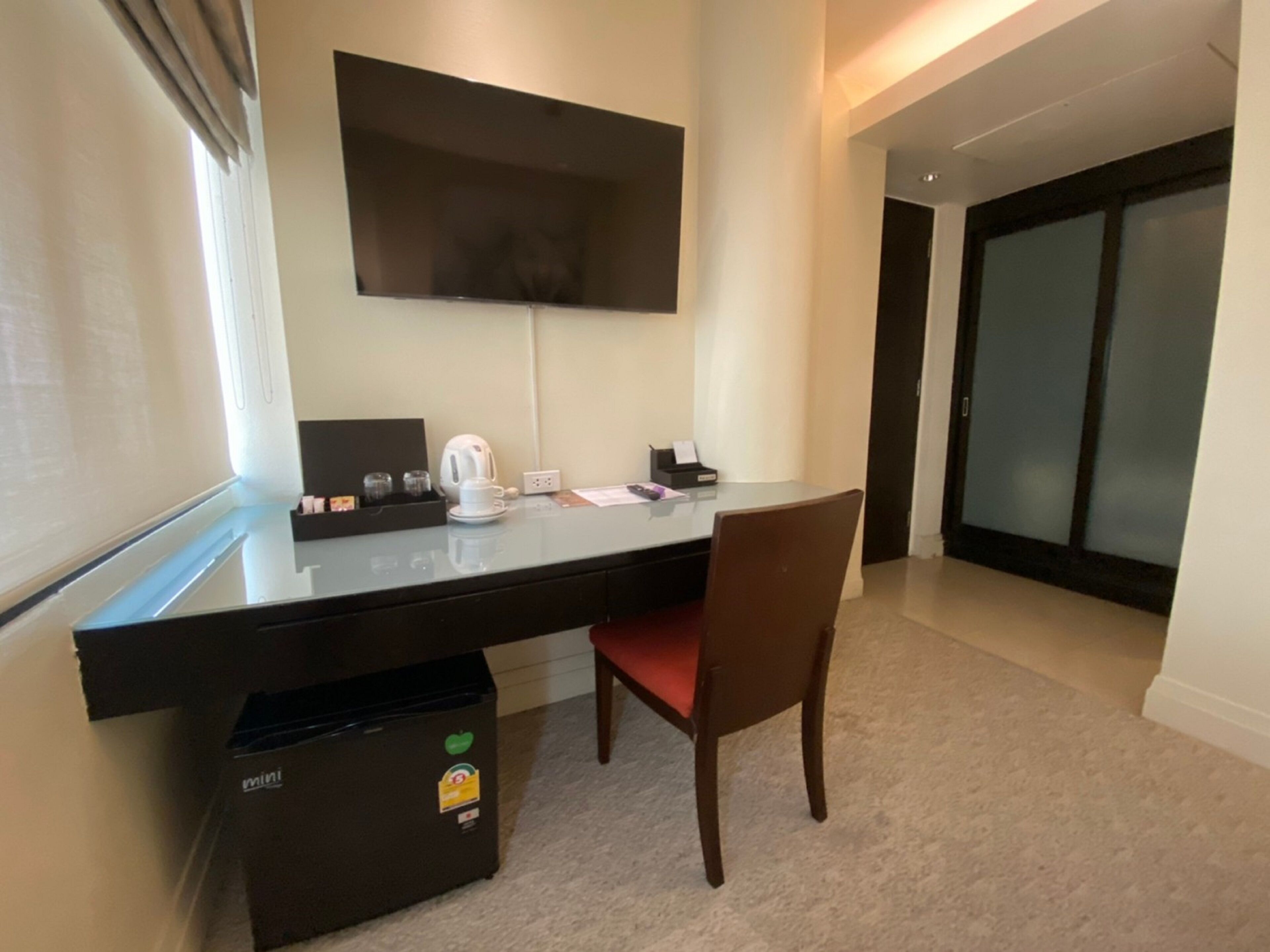 A modern hotel room with a large flat-screen TV mounted on the wall, a desk with a chair, a mini fridge, and a small table with a kettle and cups.