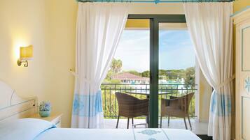 Habitación de hotel con cama, mesita de noche, lámpara de pared, ventana con cortinas, balcón con sillas de mimbre y vistas a un edificio y vegetación.