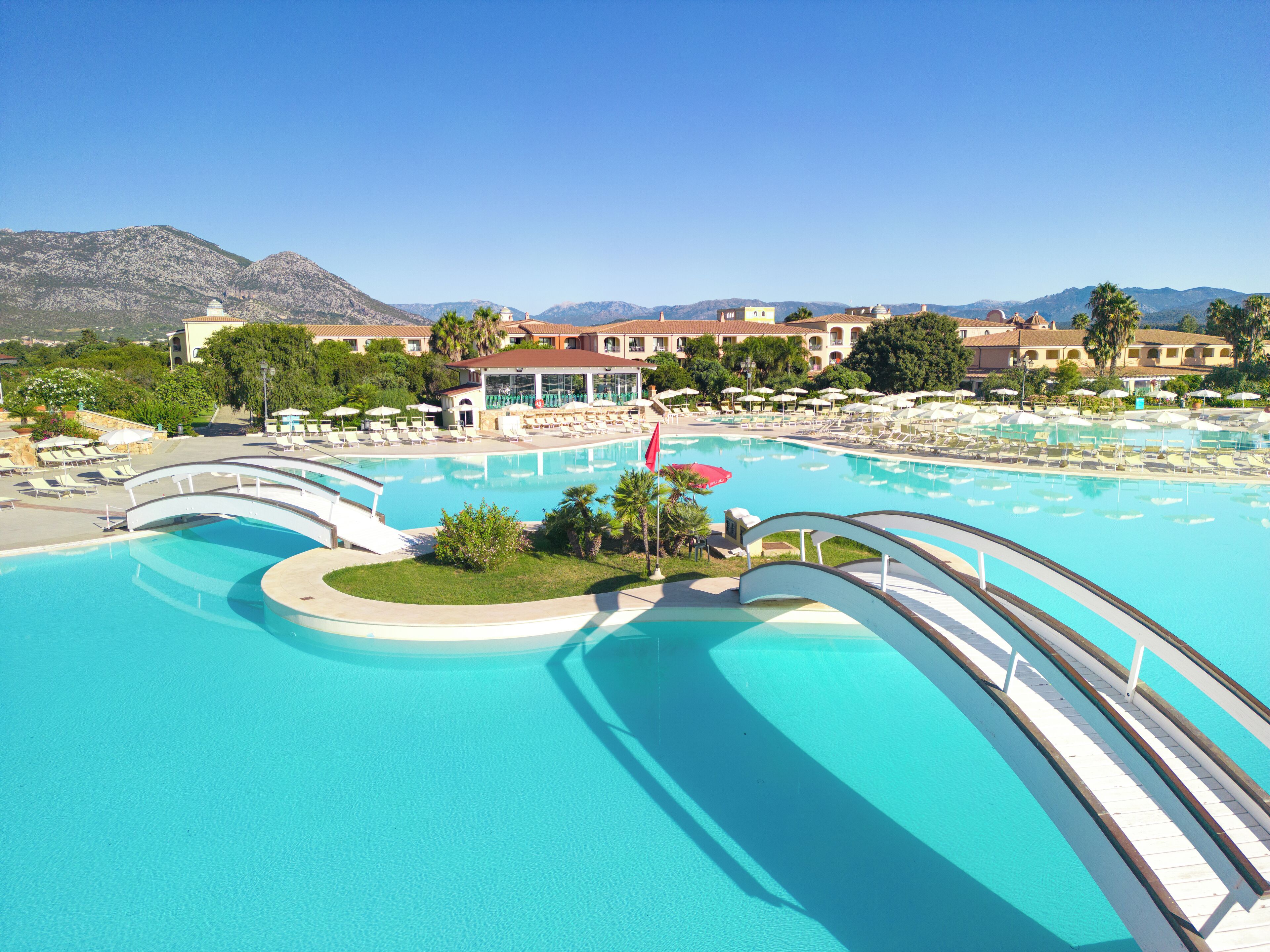 Una gran piscina con varios niveles y un puente, rodeada de sillas de descanso y sombrillas.