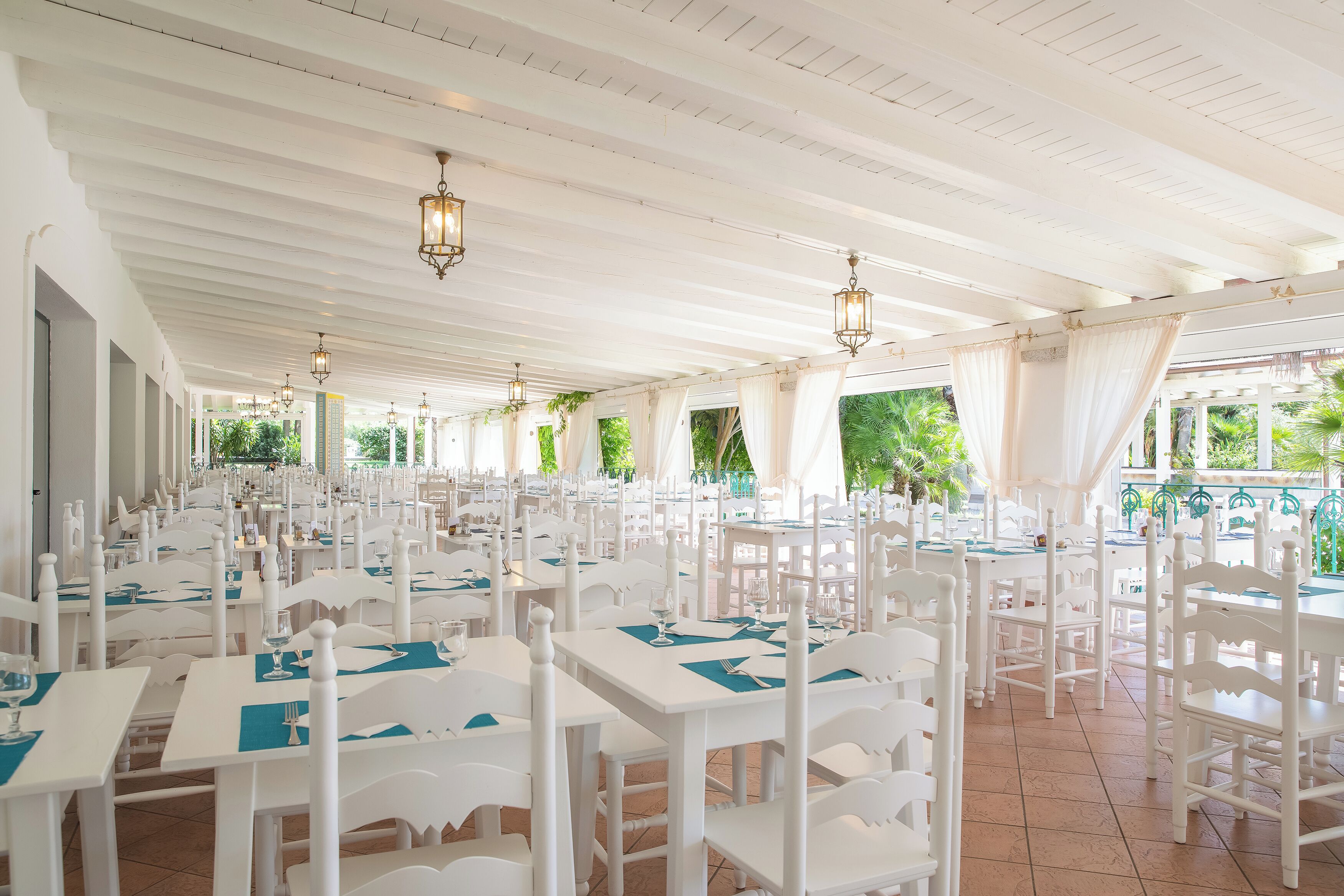 A spacious outdoor dining area with white tables and chairs, arranged in rows under a white ceiling with hanging lanterns.