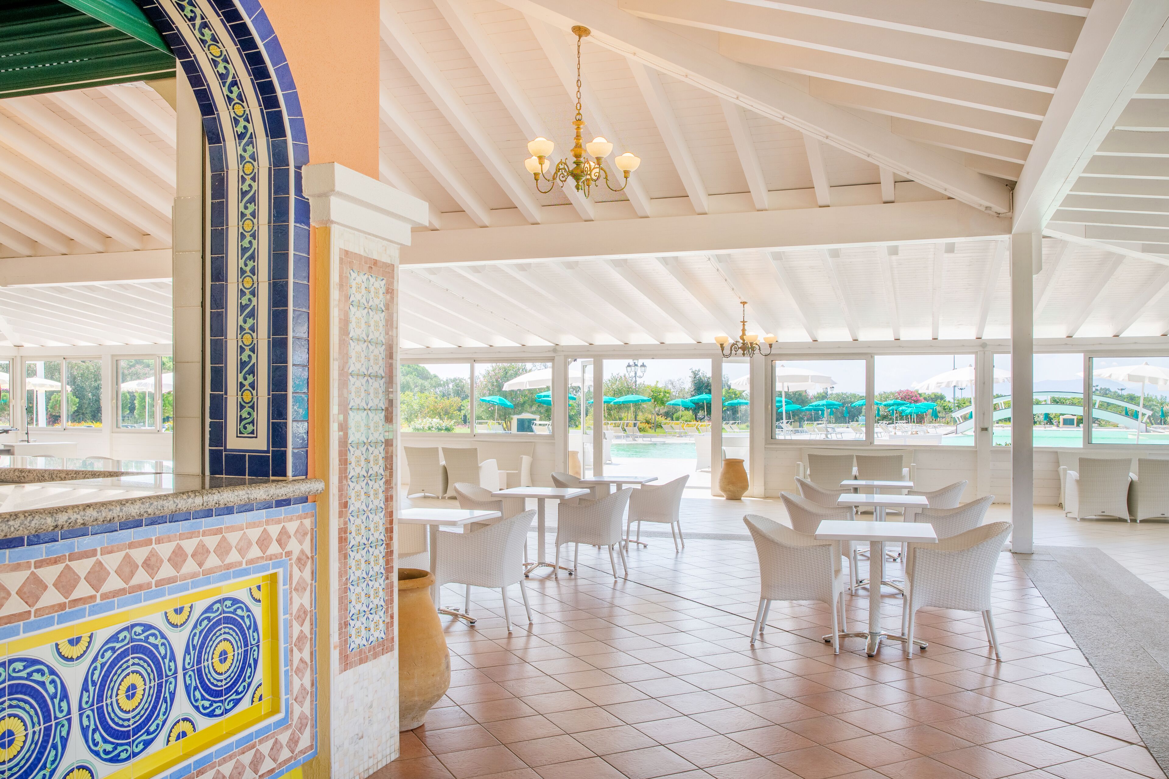 A spacious indoor area with white tiled flooring, white furniture, and large windows offering a view of the outdoors.