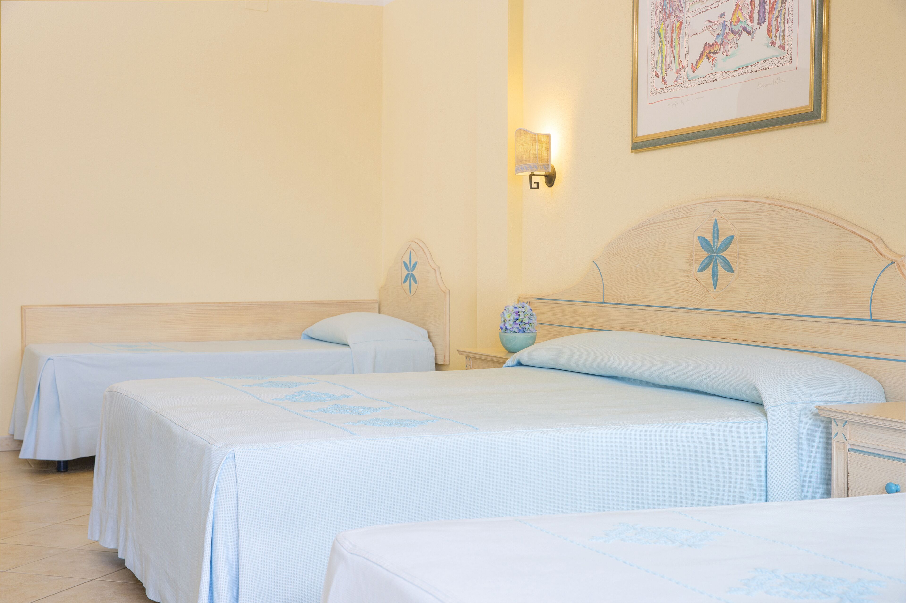 Habitación de hotel con tres camas individuales, cada una con cabecero de madera y cubrecama azul claro. Hay una lámpara de pared sobre las camas y una obra de arte enmarcada en la pared.