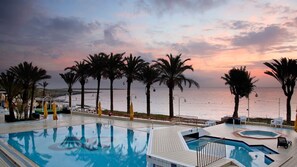 A pool area with palm trees, a view of the sea, and a sunset. - QAWRA Palace Resort & SPA (St. Paul's Bay)