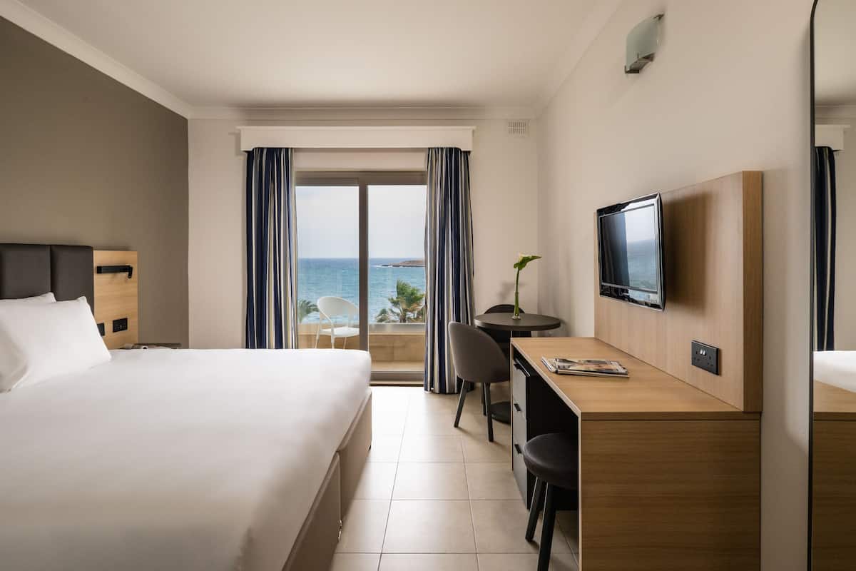 A hotel room with a bed, desk, TV, and a view of the sea.