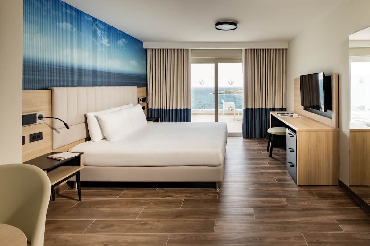 A modern hotel room with a large bed, a desk, a television, and a view of the sea.