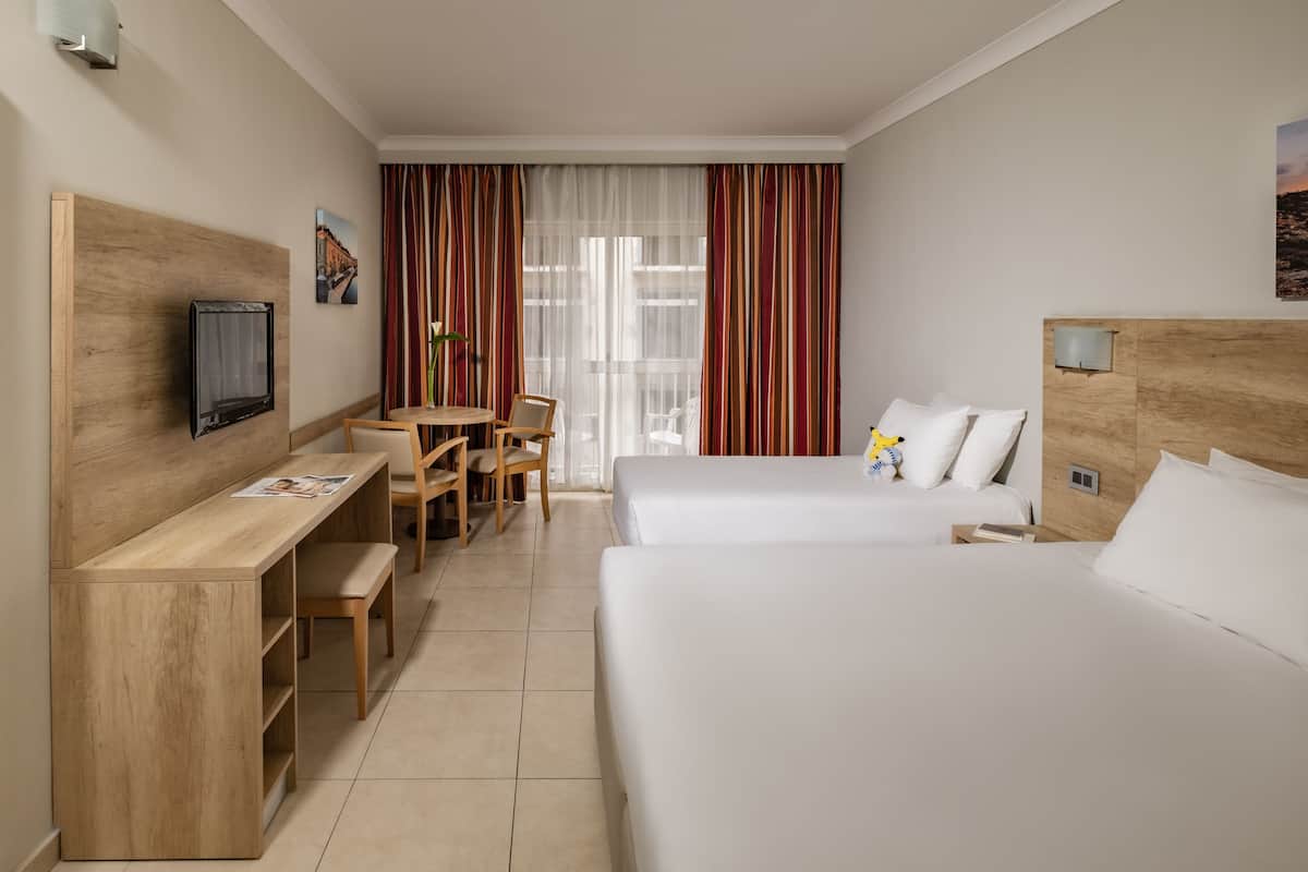 A hotel room with a wooden desk, a television, two beds, a window with curtains, and a small table with chairs.