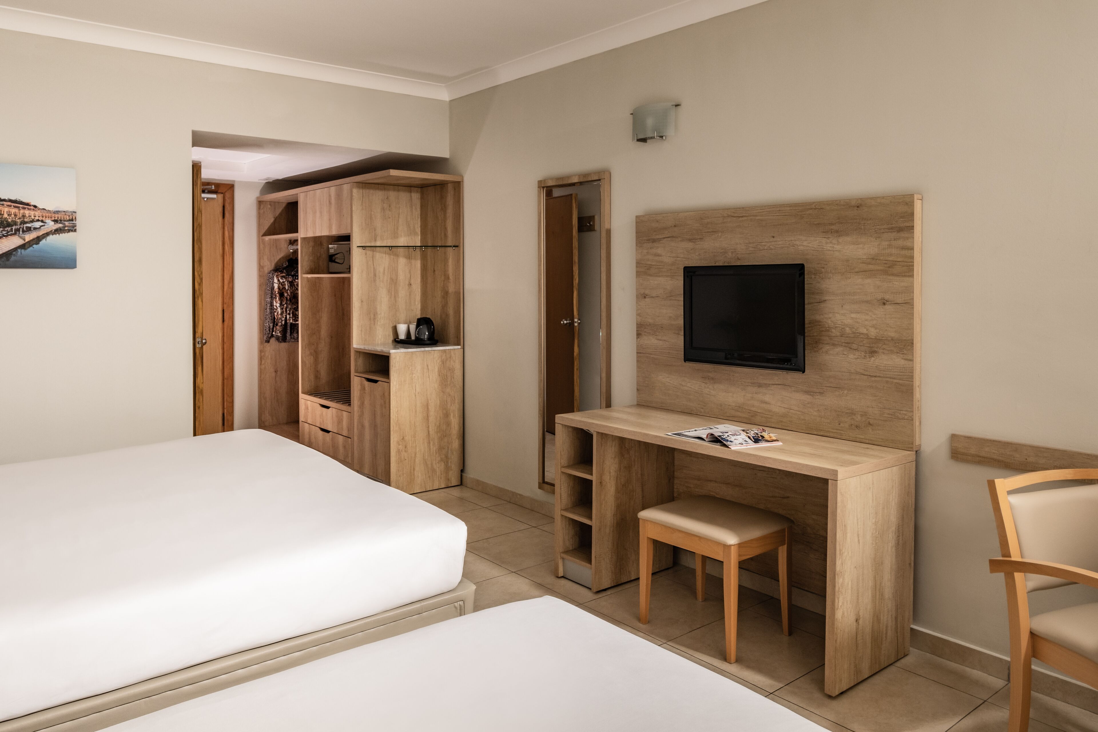 A hotel room with two beds, a wooden desk with a chair, a TV mounted on the wall, and a wardrobe.
