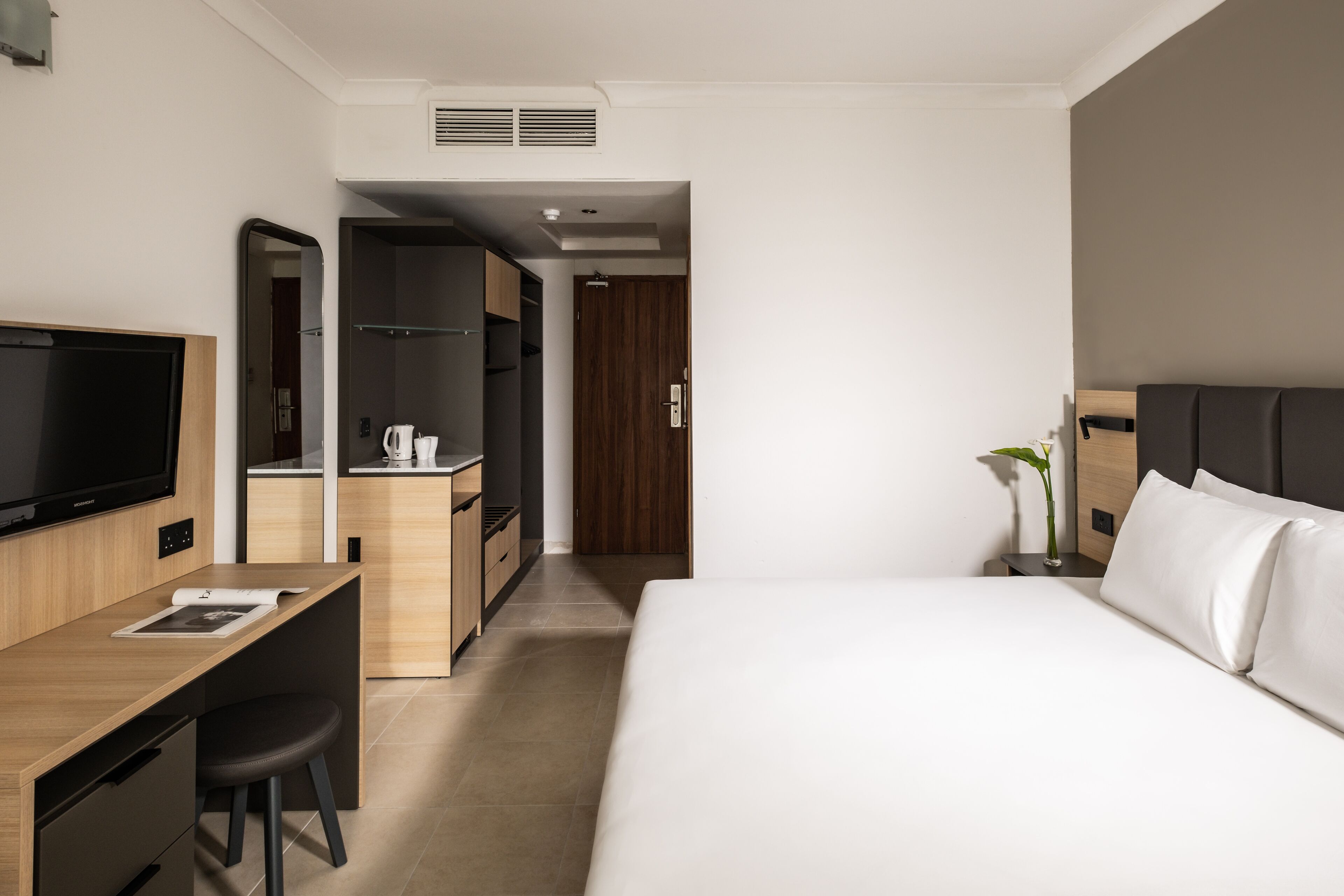 Une chambre d’hôtel moderne équipée d’un lit, d’un bureau avec une chaise, d’une télévision, d’une armoire et d’une petite kitchenette.