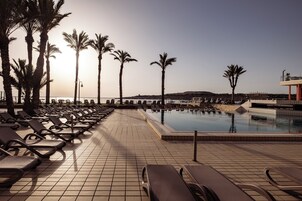 Zona junto a la piscina con tumbonas y palmeras, cielo despejado y vista al mar.