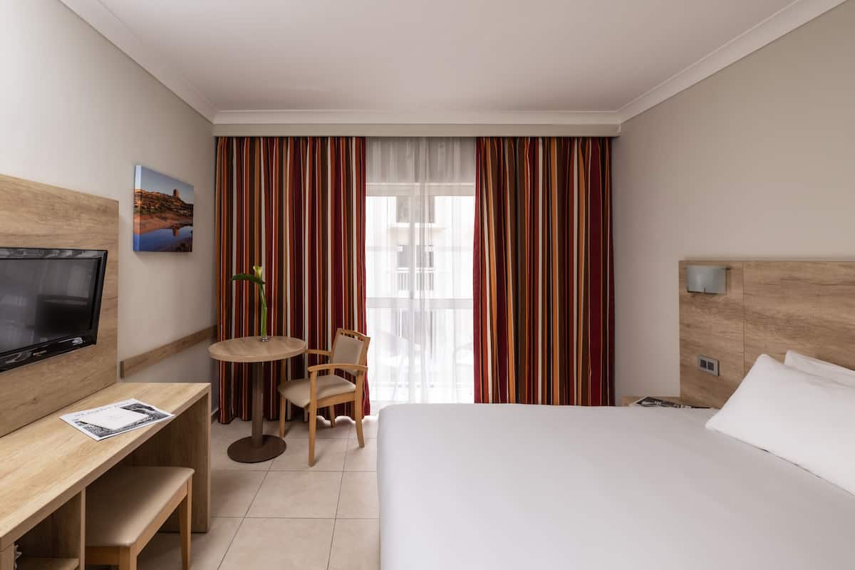 A hotel room with a bed, a desk with a chair, a television, and a window with curtains.
