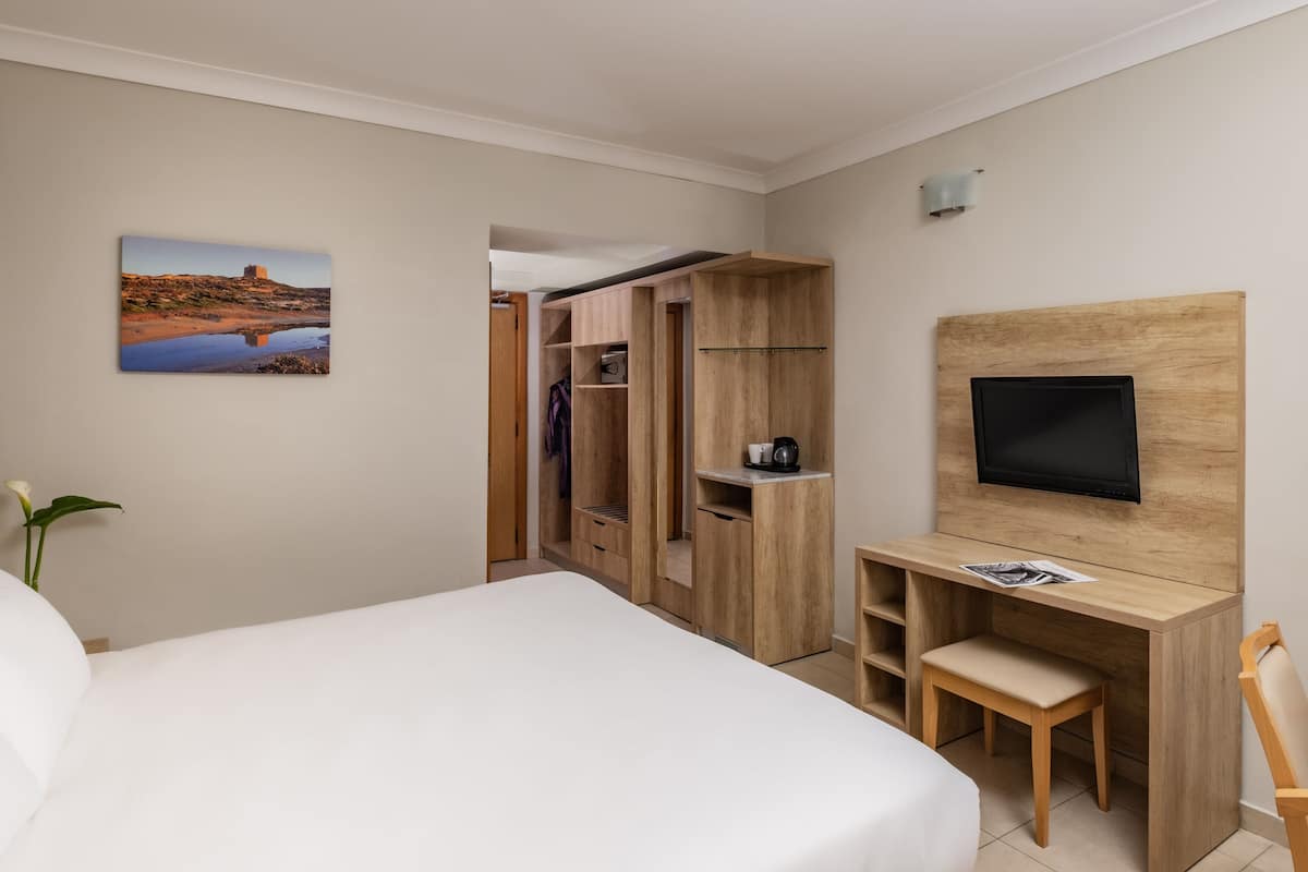 A hotel room with a bed, a desk with a chair, a TV mounted on the wall, and a wardrobe.