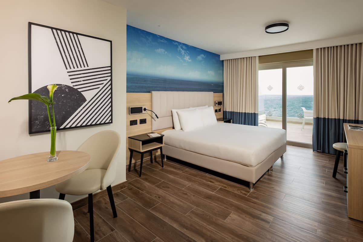 A hotel room with a bed, a desk, a chair, a small table with a vase, and a view of the sea through large windows.