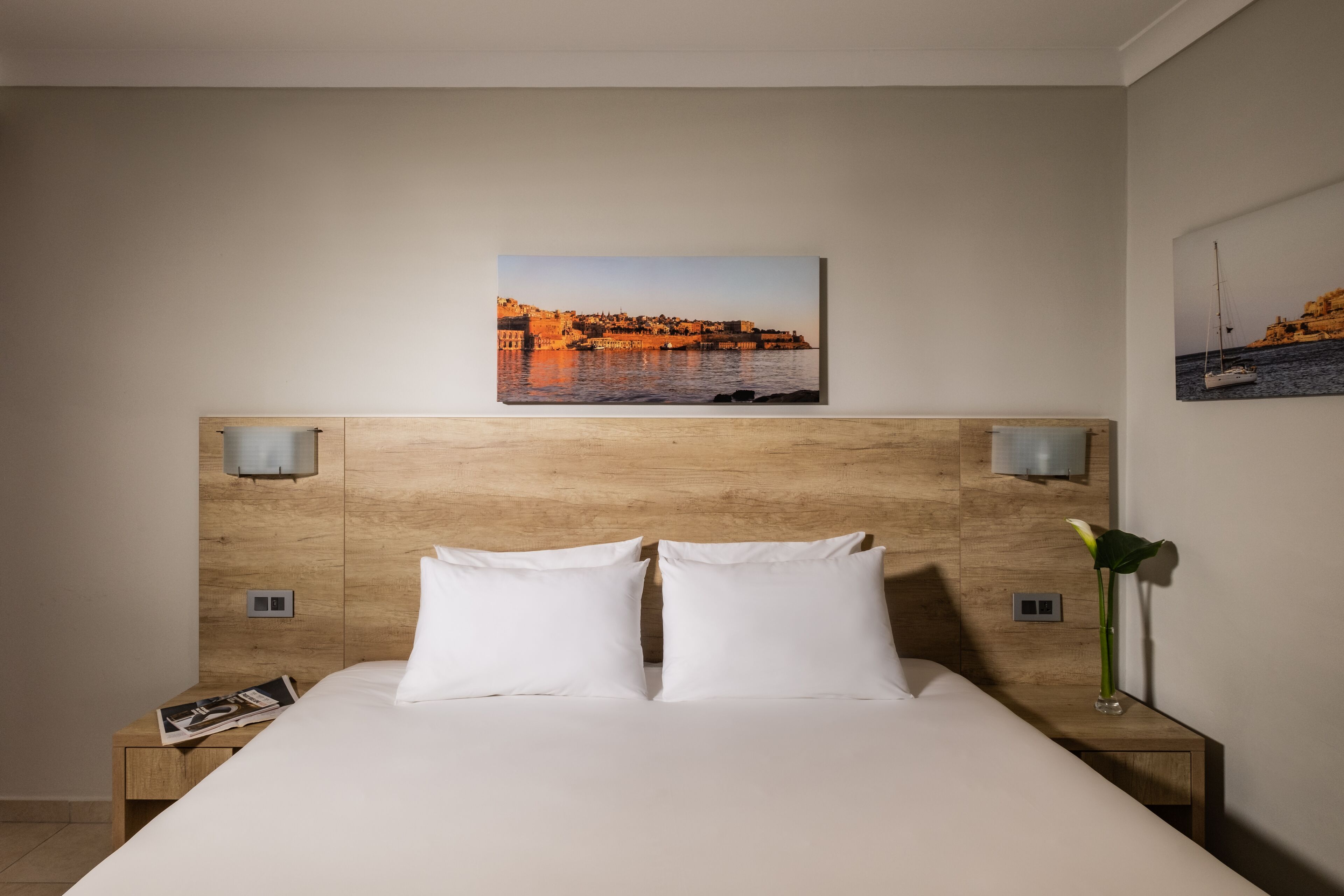 Ett hotellrum med en sänggavel i trä, två vita kuddar, ett nattduksbord med ett tidningsnummer och en tavla på väggen som föreställer en kuststad.