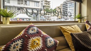 A sofa with patterned cushions and two potted plants, a window with a view of buildings and a sign, and a curtain. - Hotel Abc (Thessaloniki)