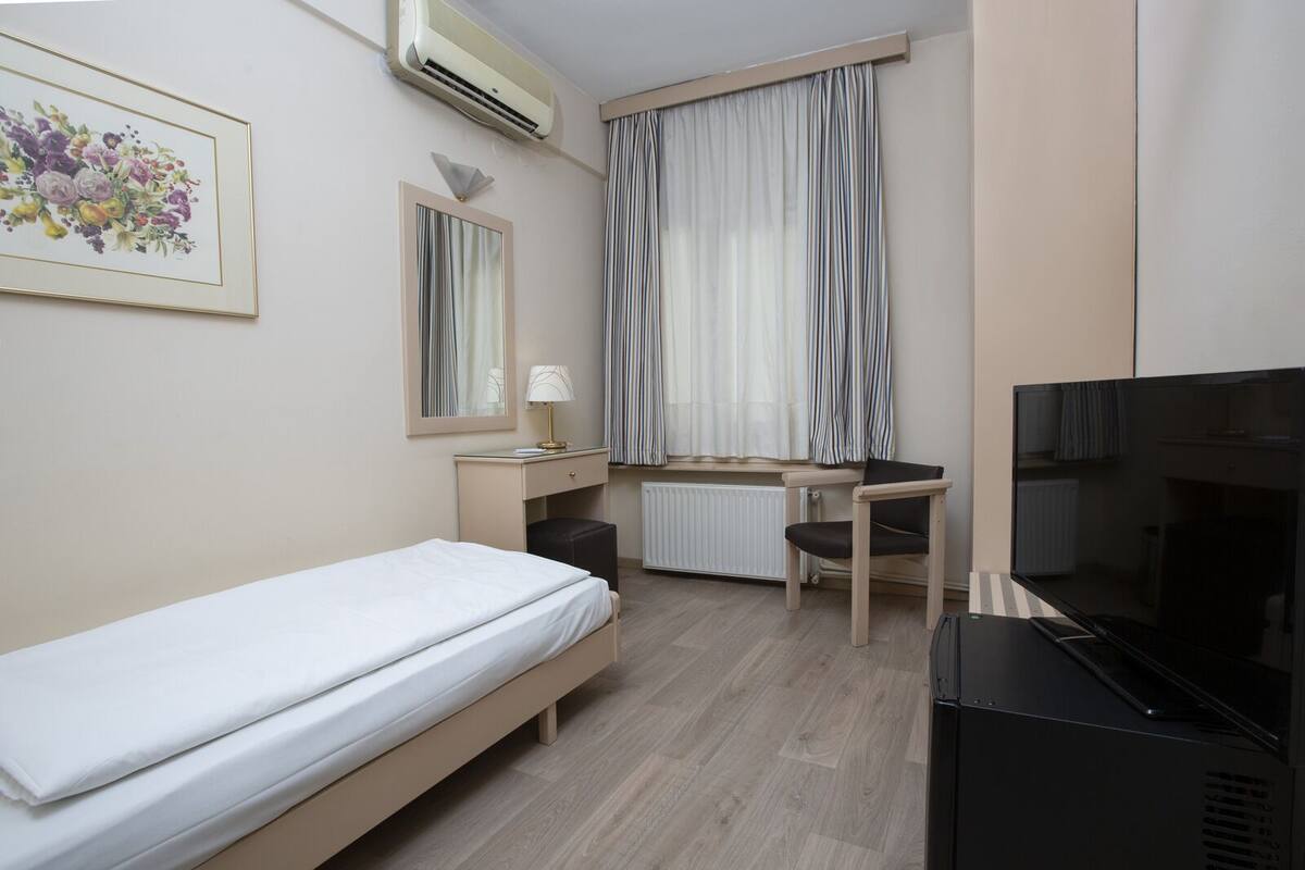 A hotel room with a bed, a desk, a chair, a television, a radiator, and a window with curtains.