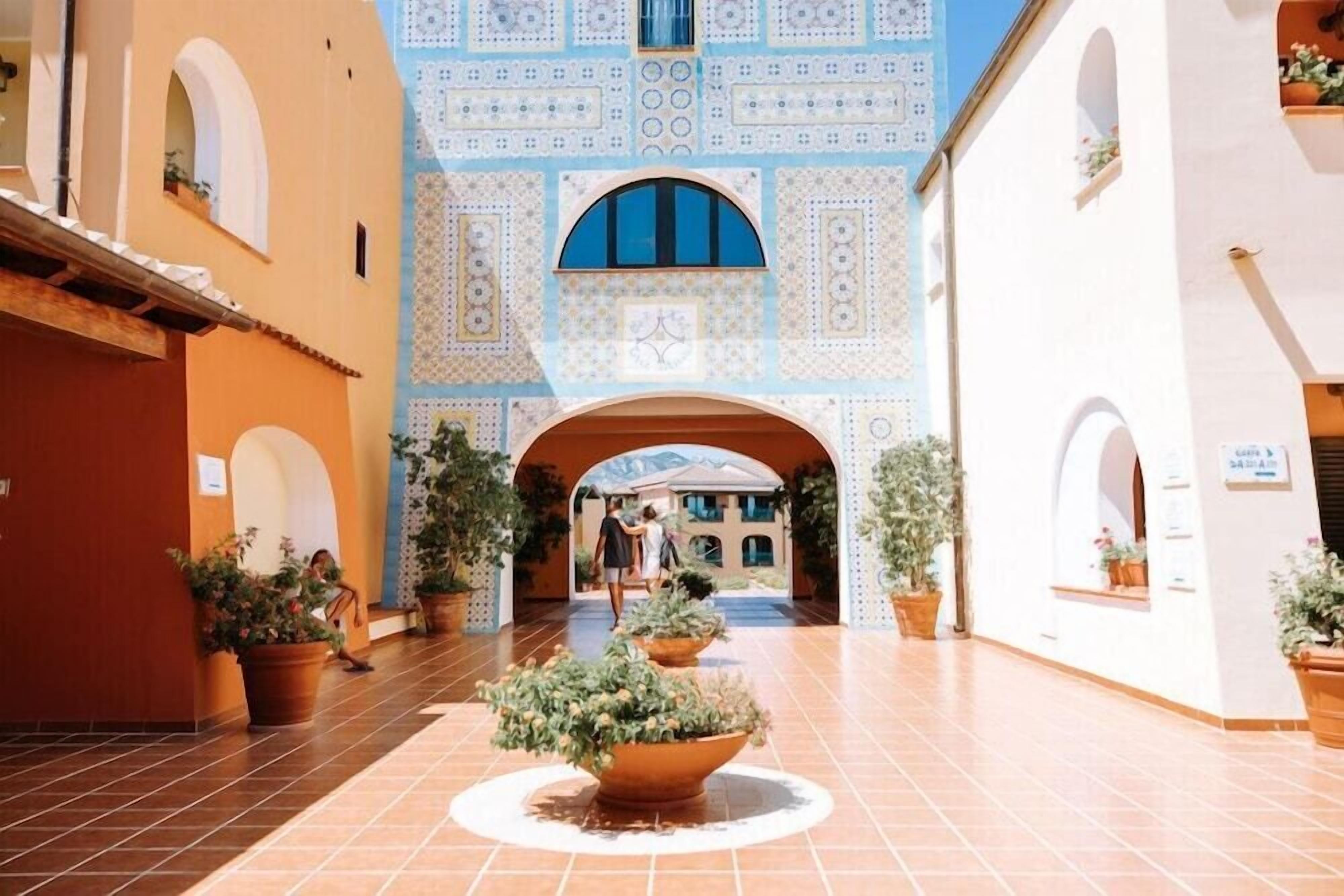 Um pátio com piso de azulejos, plantas em vasos e um edifício com paredes e arcos decorados.