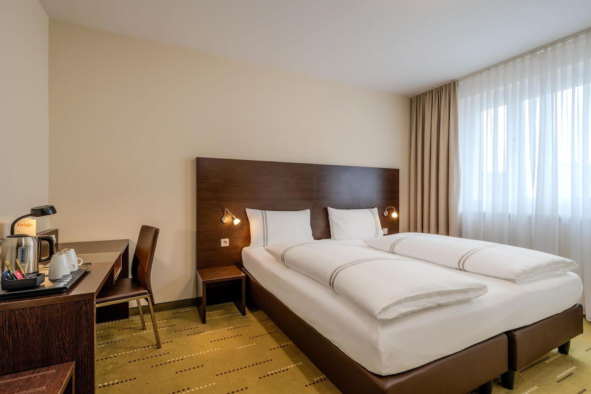 A hotel room with a large bed, a desk with a coffee maker, a chair, and a nightstand.