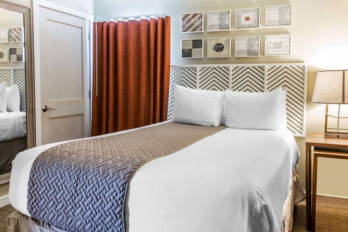 A hotel room with a bed, a nightstand, a lamp, a door, and a curtain.