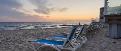 A beach with rows of sun loungers and a building with a balcony overlooking the ocean.