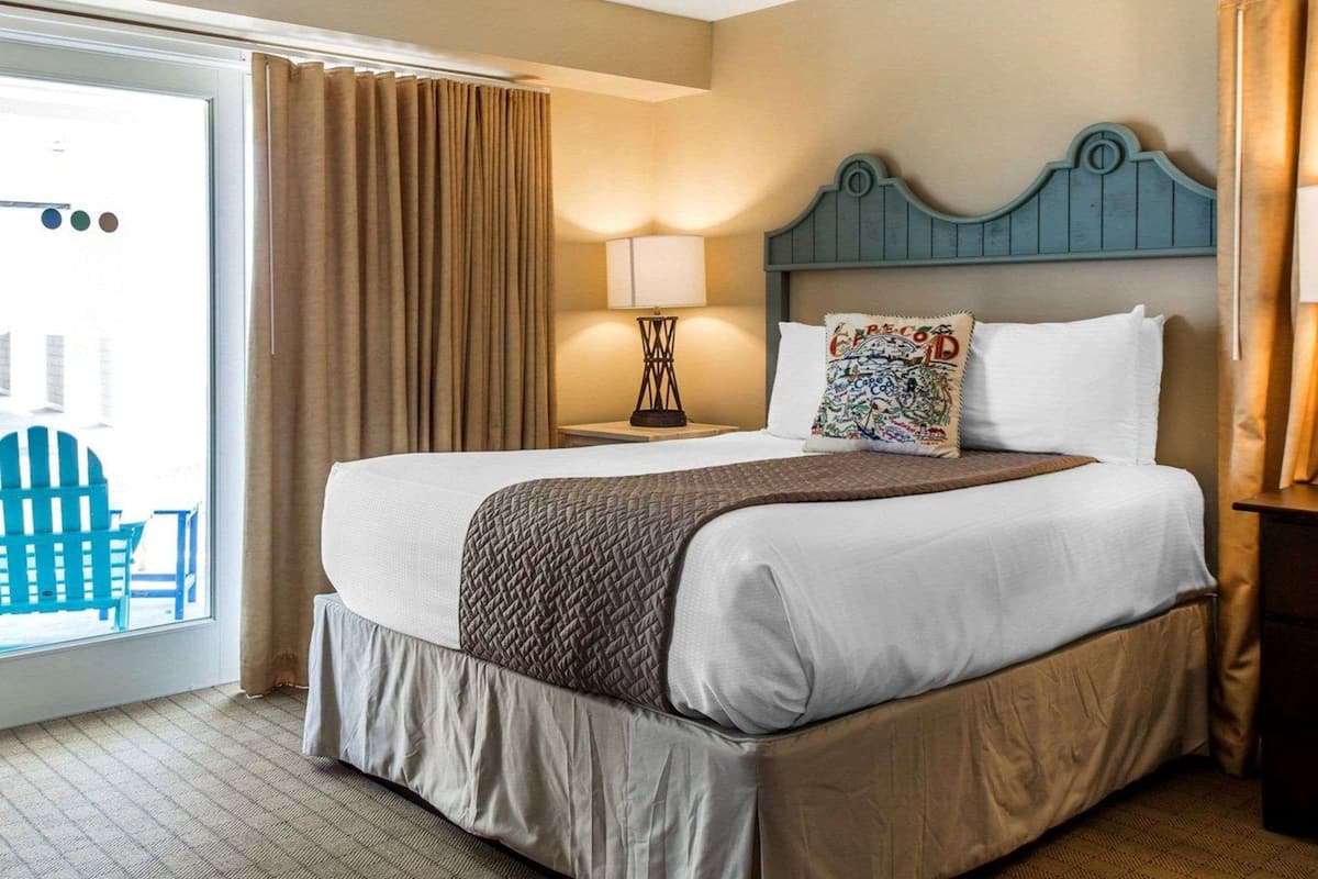A hotel room with a large bed, a headboard, a bedside table with a lamp, a window with curtains, and a view of the outside.