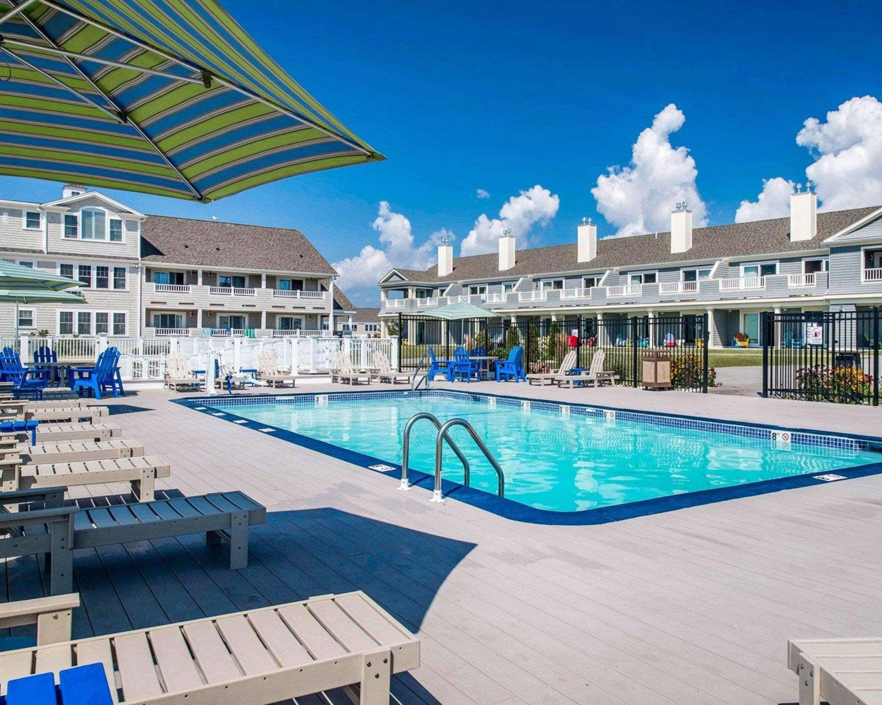 A swimming pool with lounge chairs and umbrellas, surrounded by a building with multiple balconies and a clear sky.