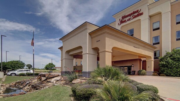 A Hampton Inn & Suites hotel with a covered entrance, a flag, and a landscaped garden.