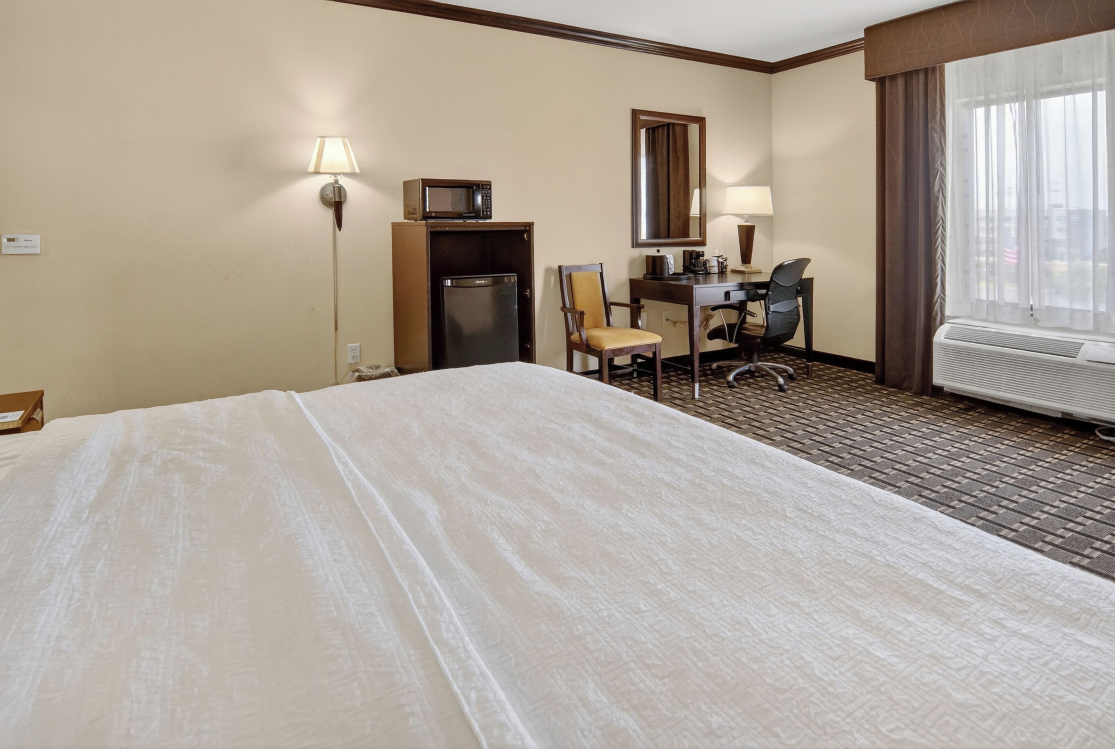 Habitación de hotel con cama, escritorio, silla, microondas, refrigerador y una ventana grande.