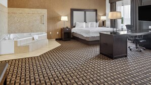 Habitación de hotel con una cama grande, un escritorio, una silla, un televisor, una lámpara de escritorio, una mesita de noche, una lámpara, una bañera y una alfombra estampada.