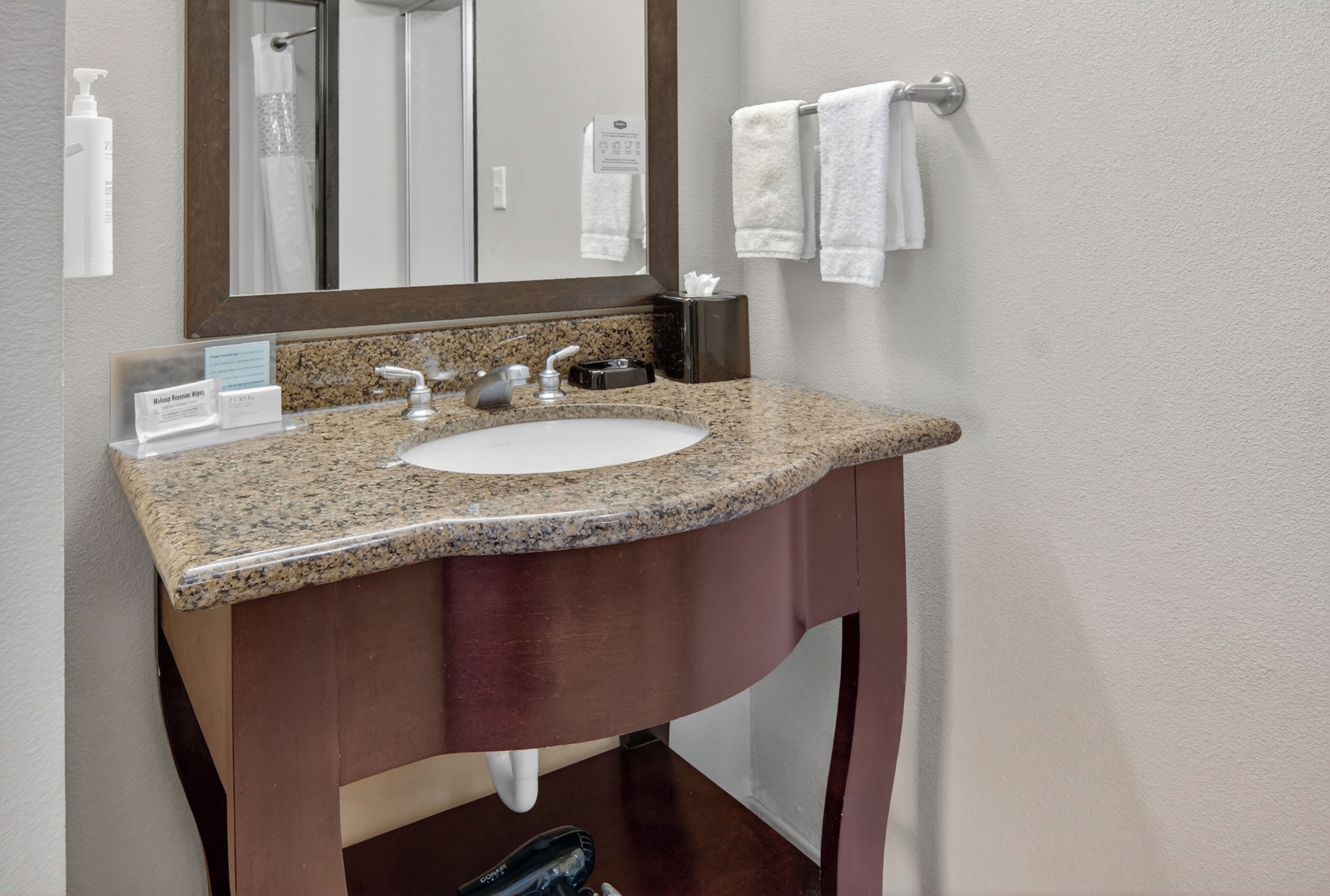 A bathroom with a granite countertop sink, a mirror, a towel rack, and a sign indicating amenities.