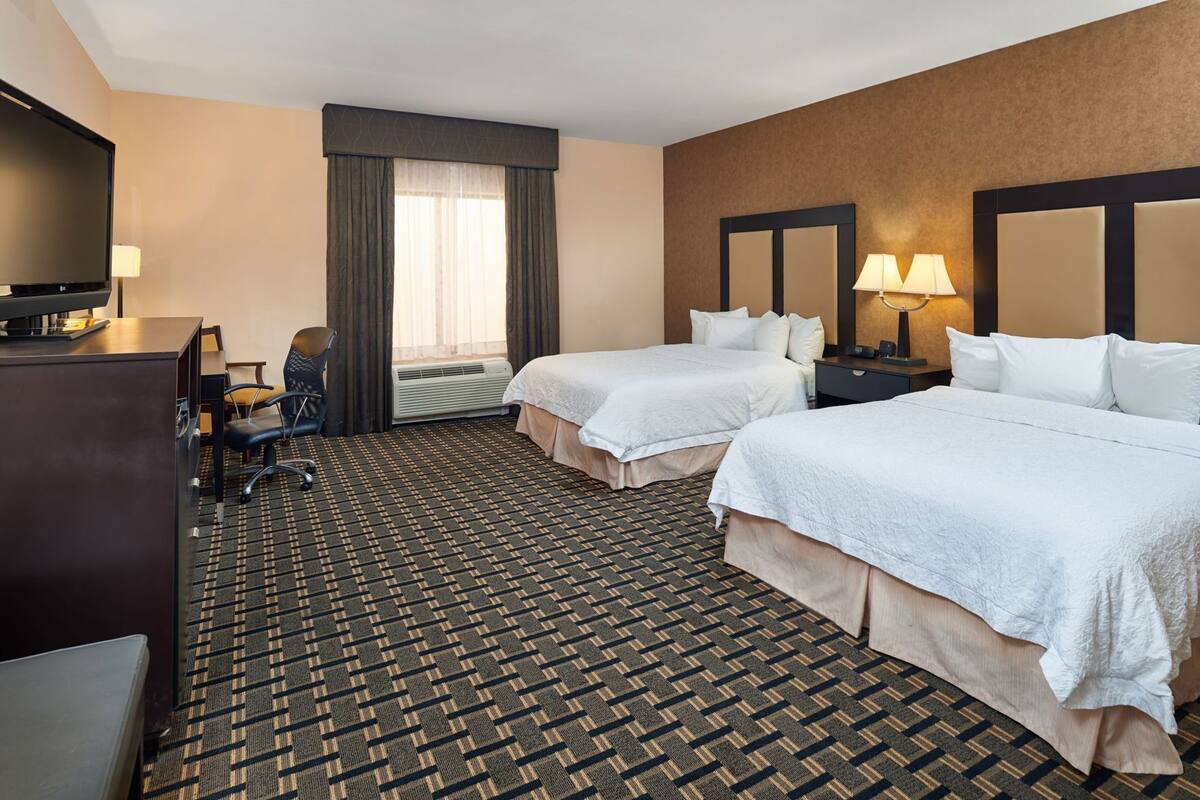 A hotel room with two beds, a desk with a chair, a television, and a window with curtains.