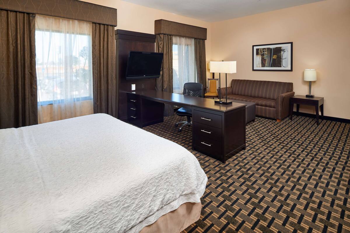 A hotel room with a large bed, a desk with a TV, a chair, a lamp, and a framed picture on the wall.