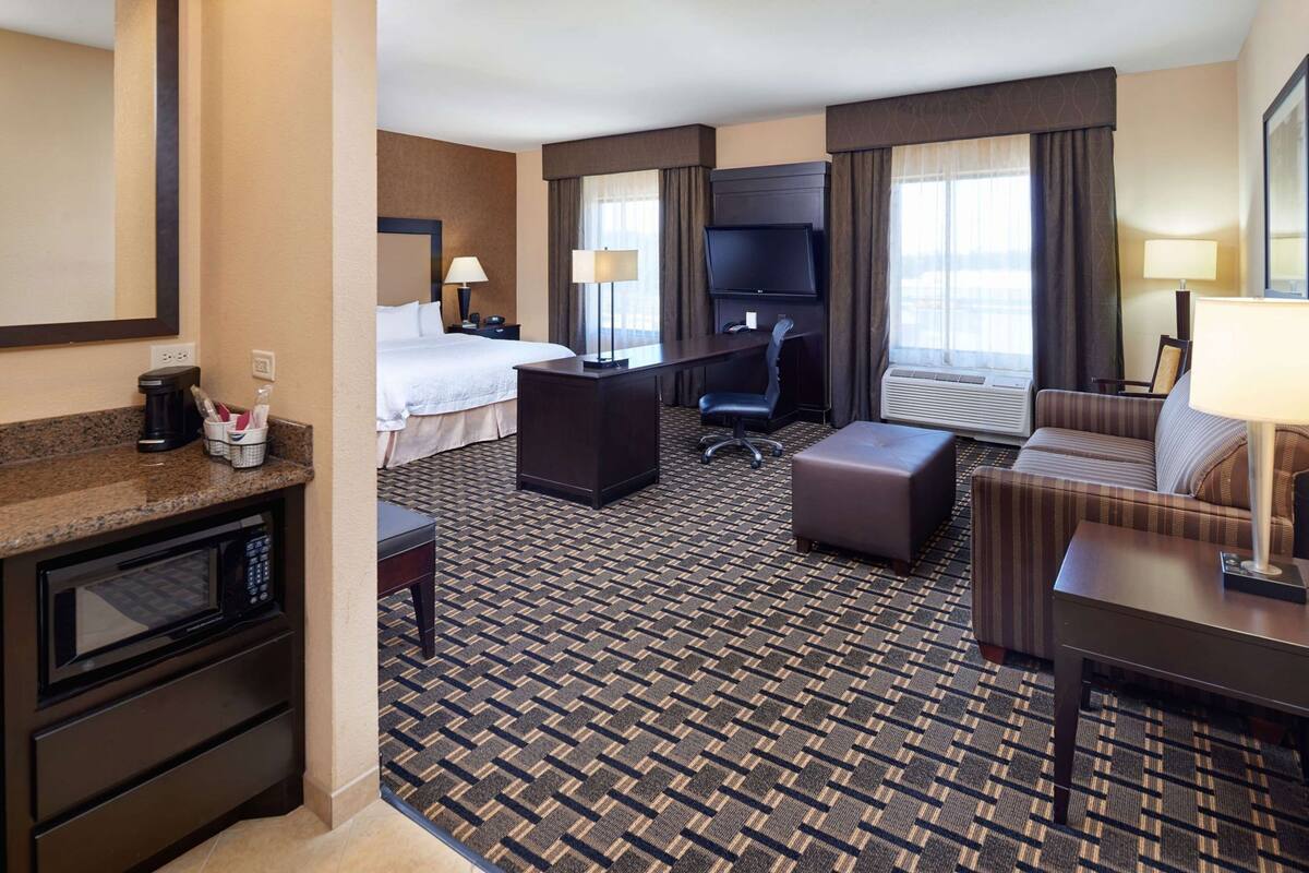 A hotel room with a bed, desk, TV, kitchenette, and a patterned carpet.