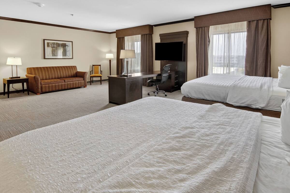 A hotel room with two beds, a desk, a chair, a sofa, a TV, and a window with curtains.