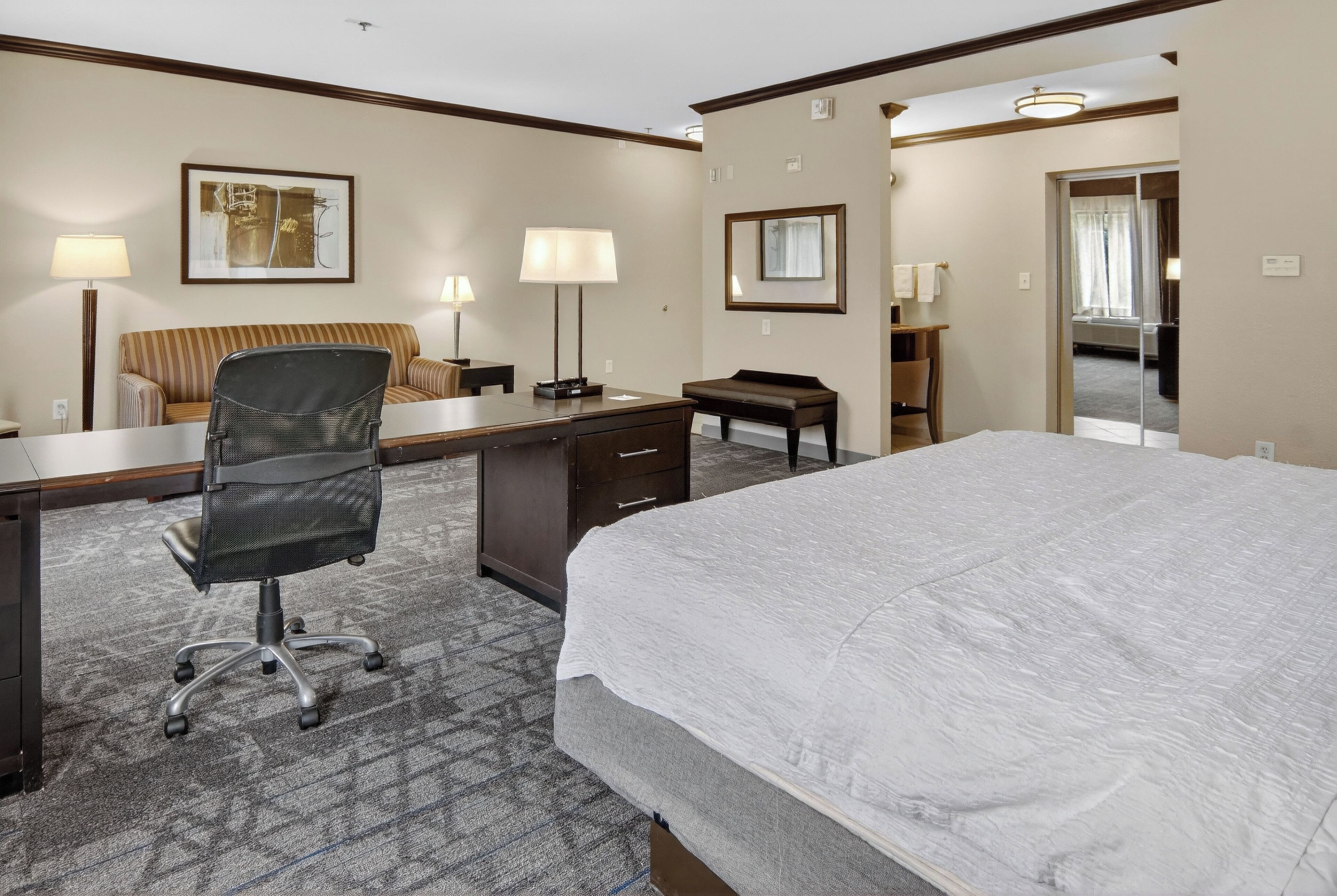 A hotel room with a bed, desk, office chair, lamps, and a mirror.