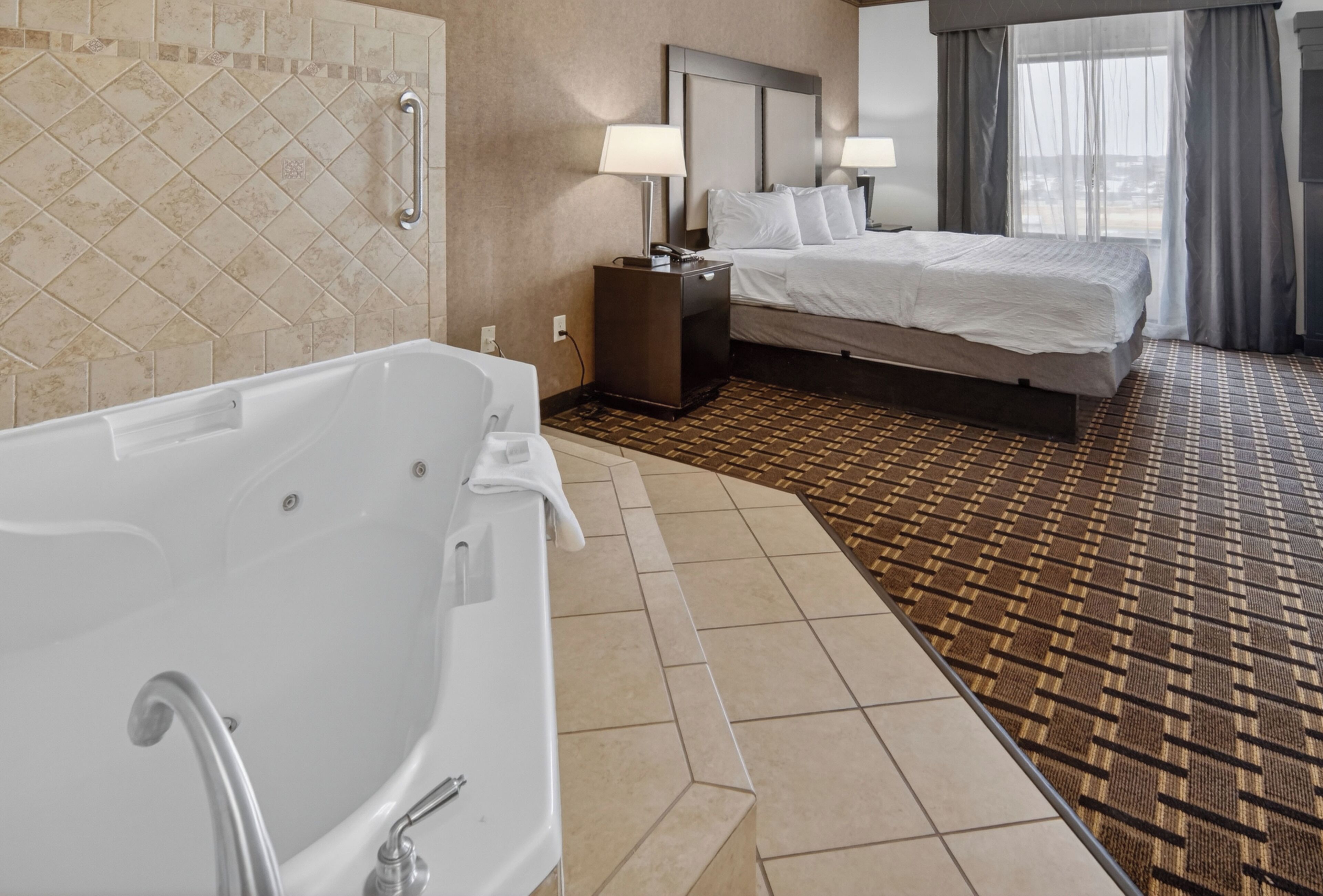 Habitación de hotel con una bañera grande, una cama con sábanas blancas, una mesita de noche con lámpara y una alfombra estampada.