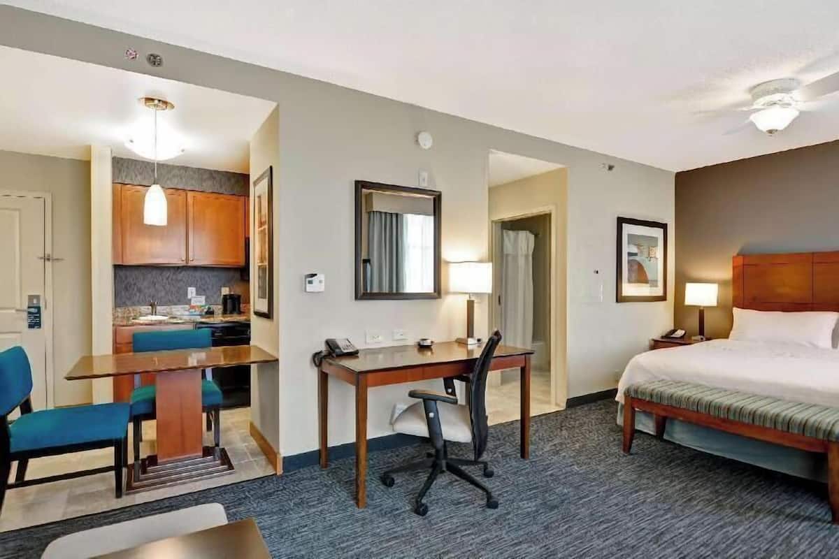 A hotel room with a bed, desk, chair, kitchenette, and a bathroom.