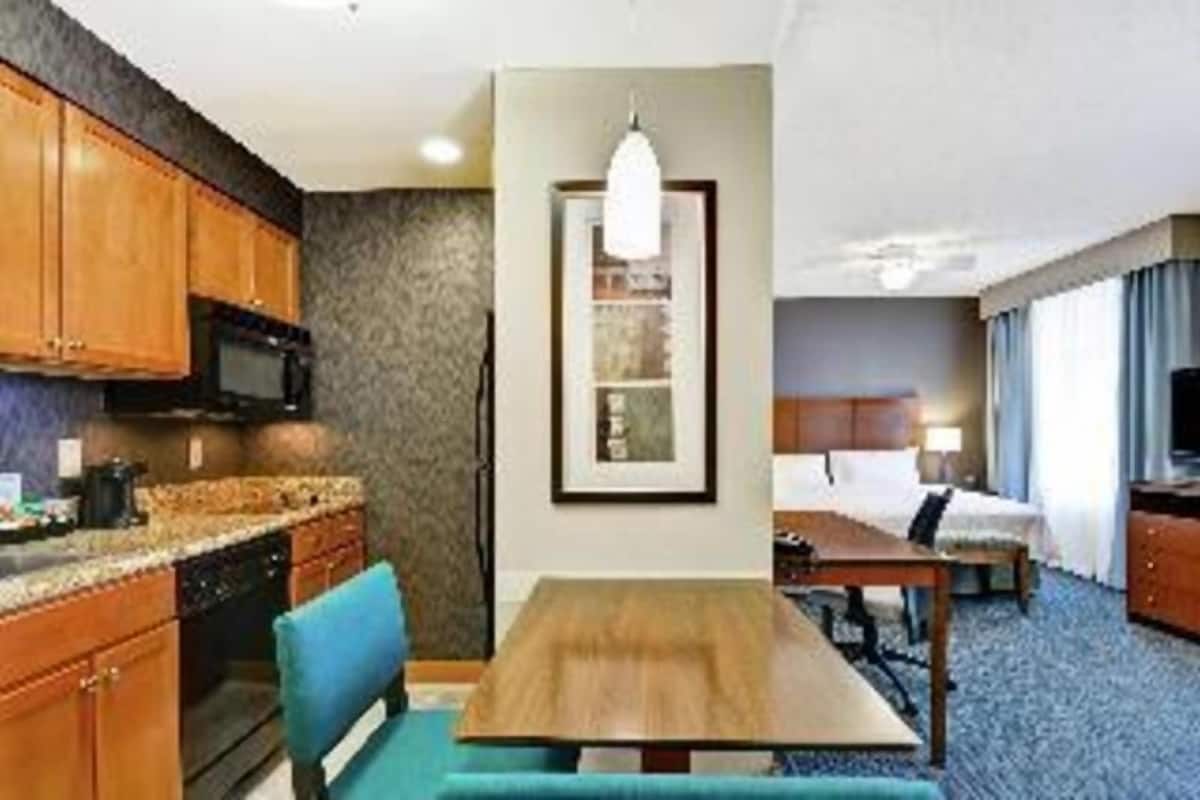 A hotel room with a kitchenette, a dining area, a bed, and a large window.