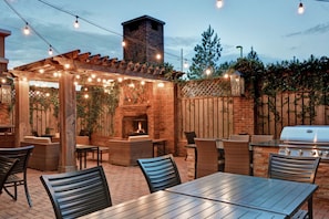 Un patio al aire libre con suelo de ladrillo, pérgola de madera, luces colgantes y mobiliario exterior.