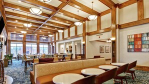 A spacious lobby with high ceilings, wooden beams, and a mix of seating areas including a long bench and round tables.