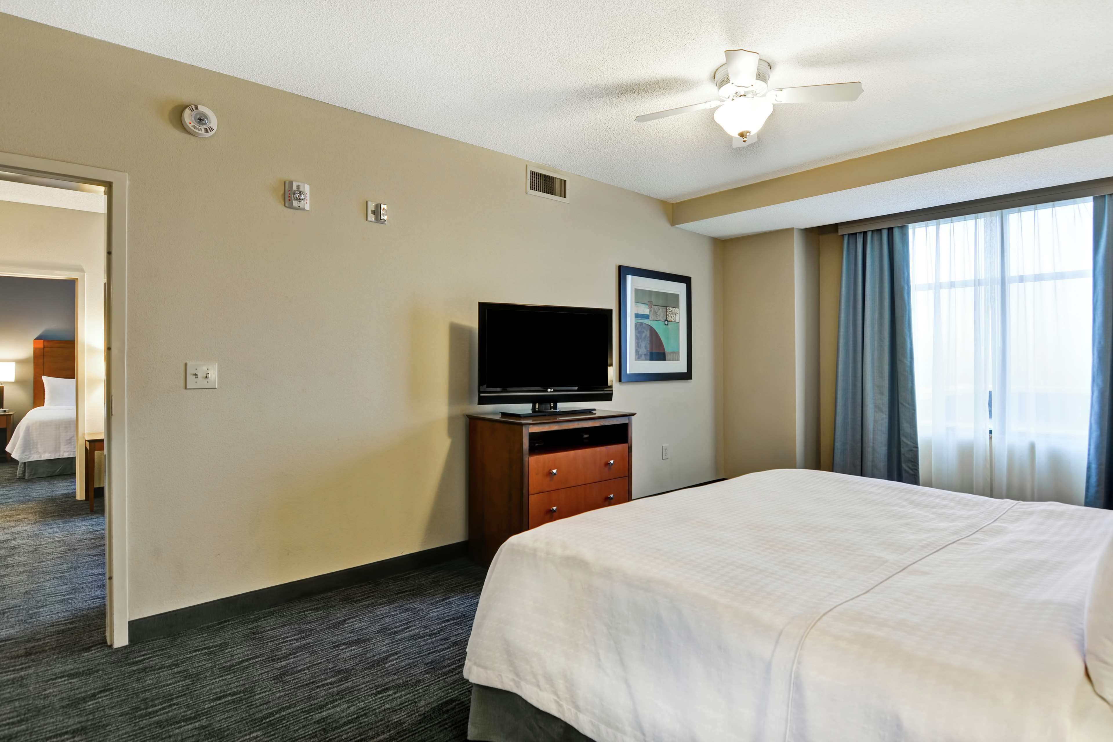 Ein Hotelzimmer mit einem Bett, einem Fernseher auf einer Holzkommode, einem Deckenventilator und einem Fenster mit Vorhängen.