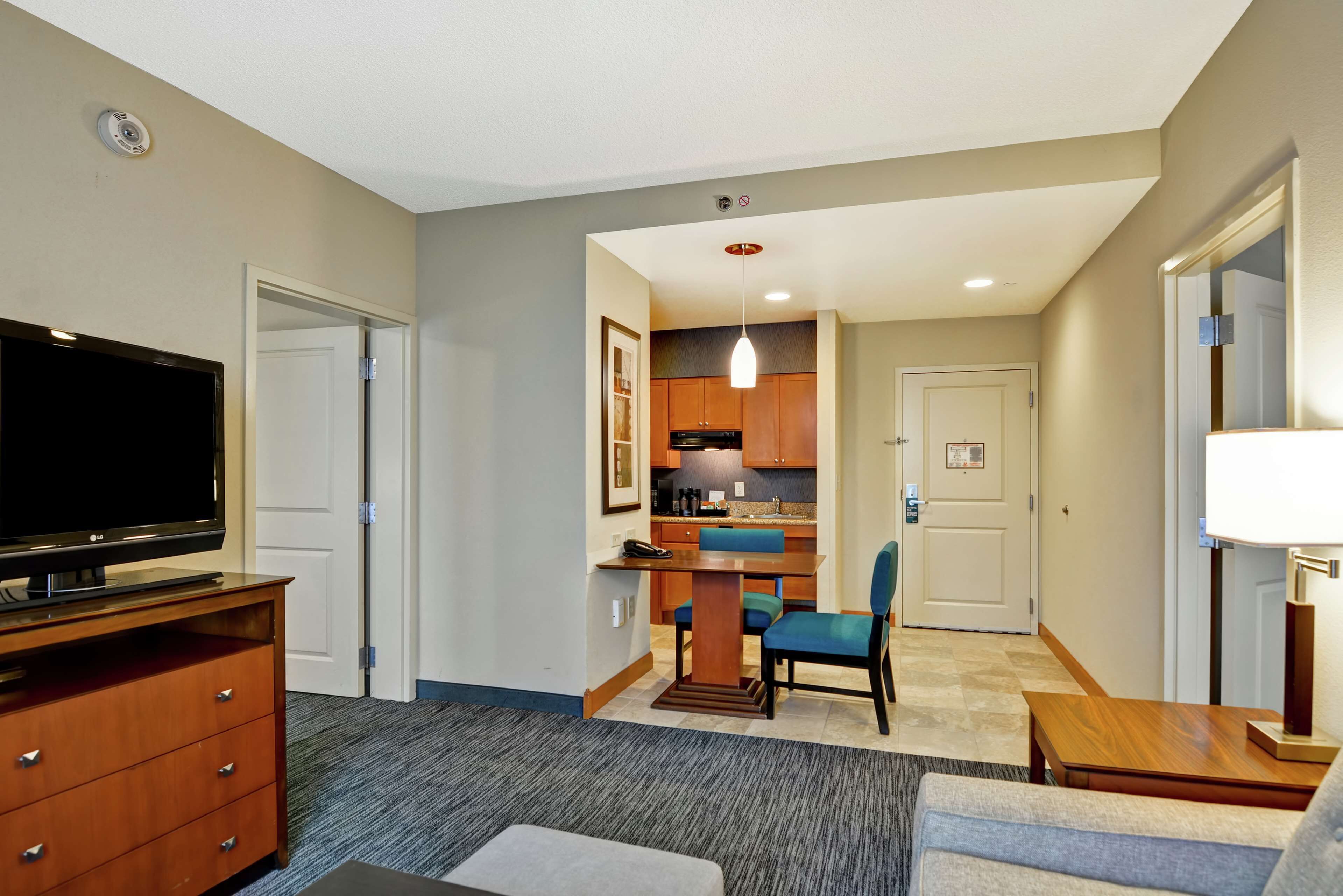 A hotel room with a flat-screen TV, a wooden dresser, a dining table with chairs, a kitchen area, and a lamp.