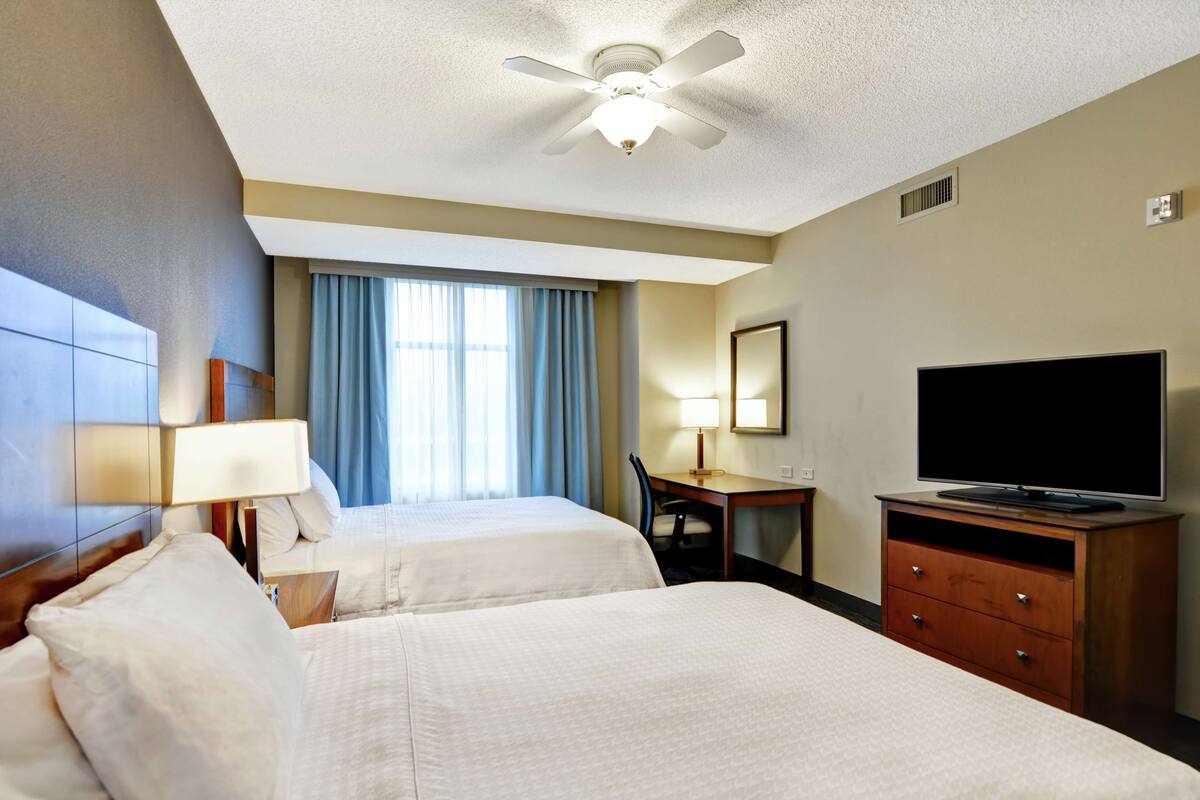 A hotel room with two beds, a desk with a chair, a television, and a ceiling fan.