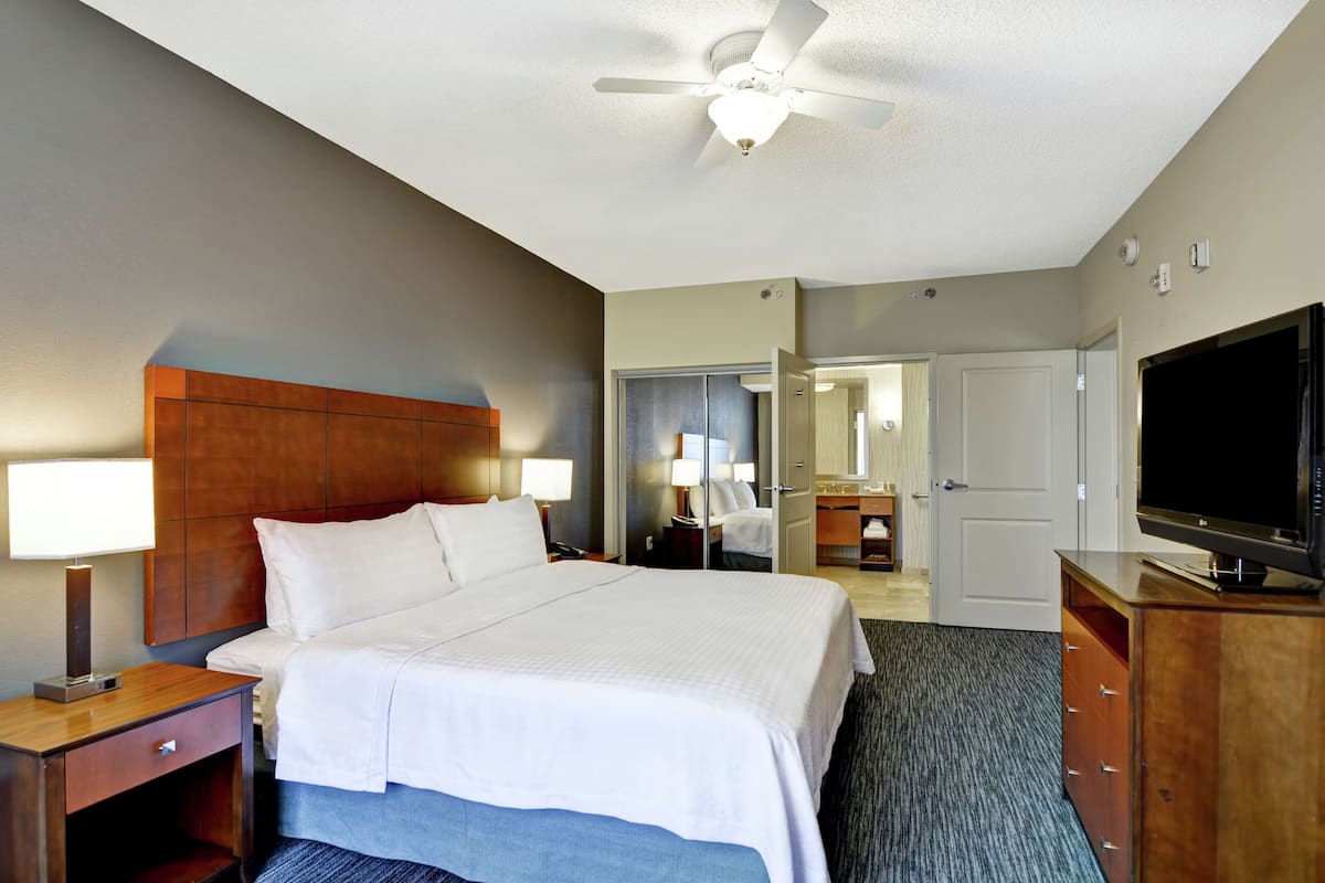 A hotel room with a large bed, a ceiling fan, a TV, and a wooden nightstand with a lamp.