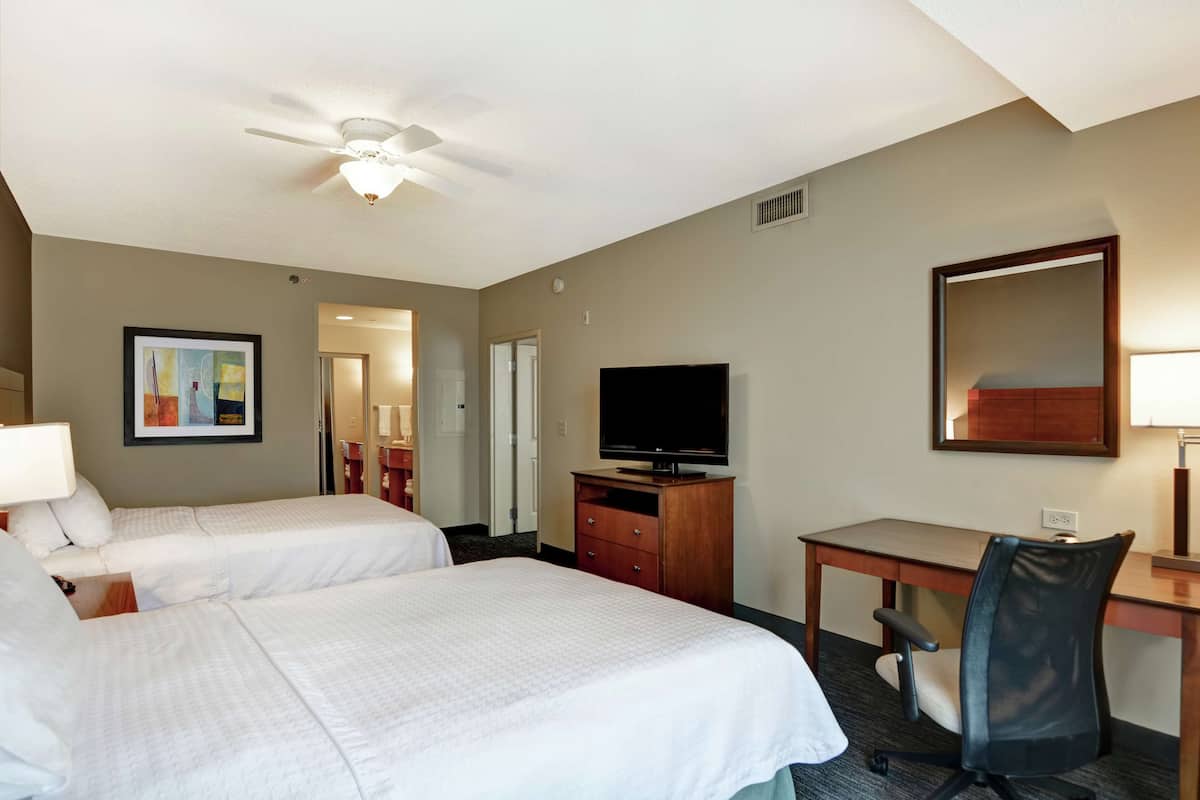 A hotel room with two beds, a desk, a chair, a TV, and a ceiling fan.