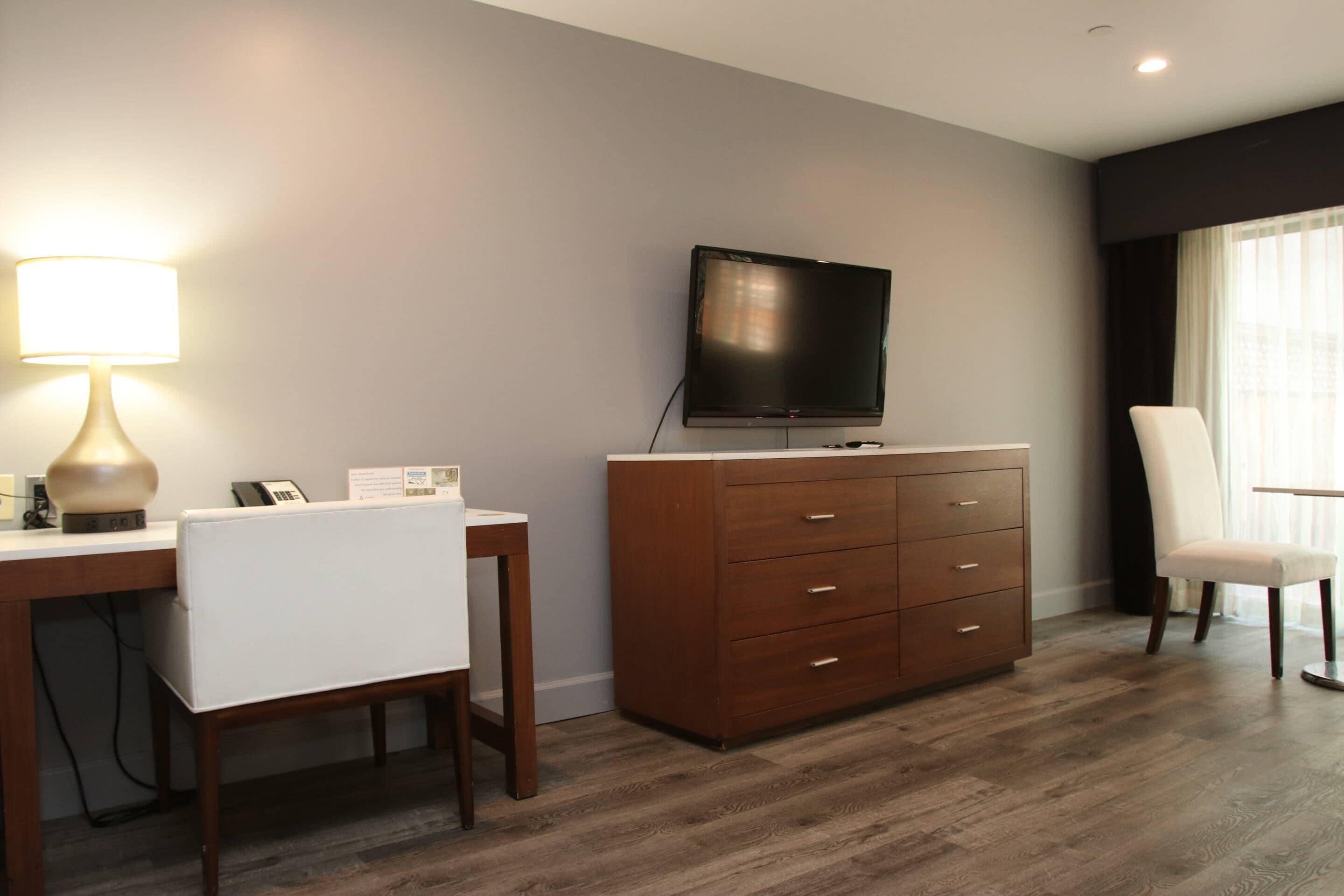 A hotel room with a wooden desk, a white chair, a brown dresser, a television, and a window with curtains.