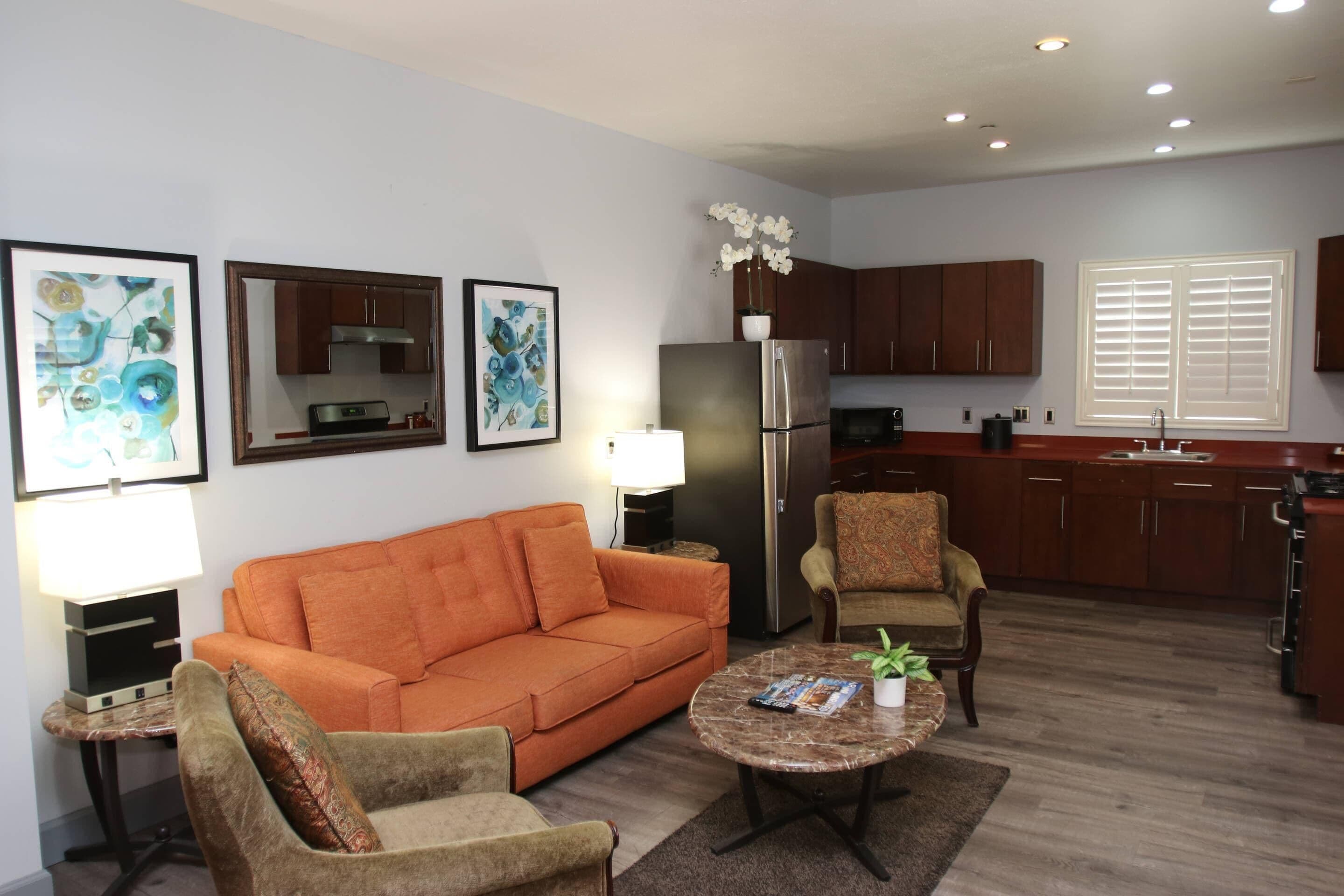 A living room with a sofa, armchair, and coffee table, a kitchen with wooden cabinets, and a window with blinds.