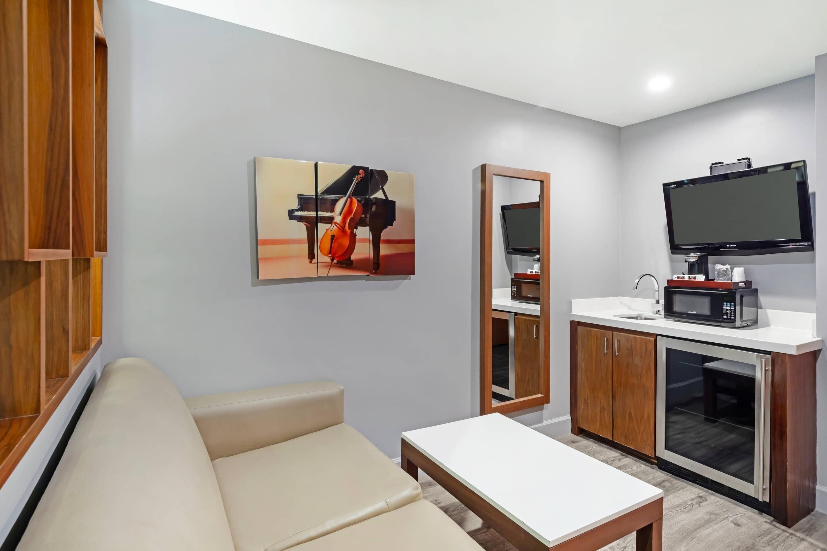 Una sala de estar moderna con un sofá, una mesita, un cuadro de un violonchelo, un televisor y una kitchenette con microondas y fregadero.