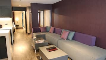 Una sala moderna con un sofá gris, una mesa de centro y una mesita auxiliar. Se observa un área de cocina al fondo con una encimera y armarios.